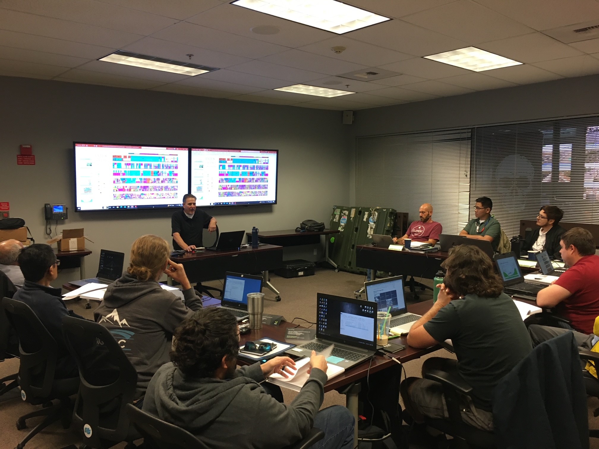Instructor Jonathan David explains a concept during the Radio Frequency Fundamentals course. A group of people sit in a semi-circle around an instructor with a slide presentation up in the background.